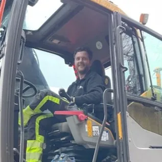 Mann med svart jakke og refleksbukse, i en gravemaskin